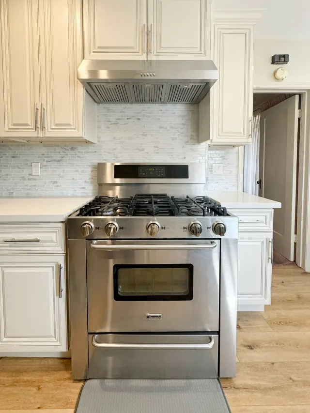 Stainless gas range and vent hood under cream raised-panel cabinets with a marble mosaic backsplash in a Riverside kitchen remodel