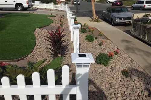 White picket fence with post lights beside a front yard of turf and rock planting